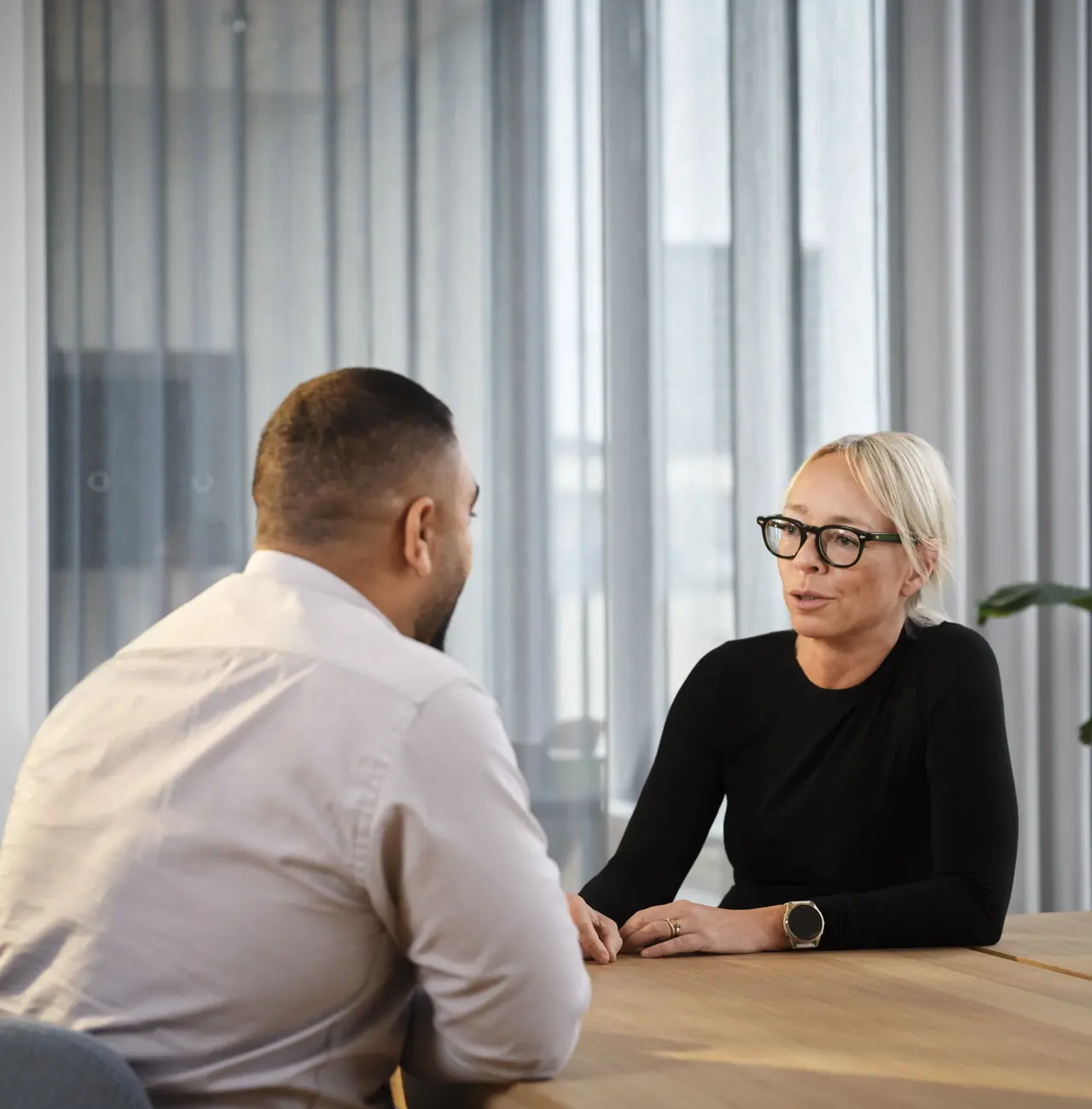 mand og kvinde sidder ved bord og taler. De er i et mødelokale. Plante i baggrund.