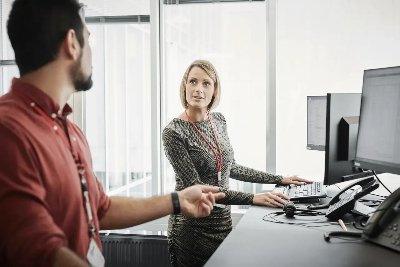 To personer står ved computer bord på et kontor. En dame og en mand. 