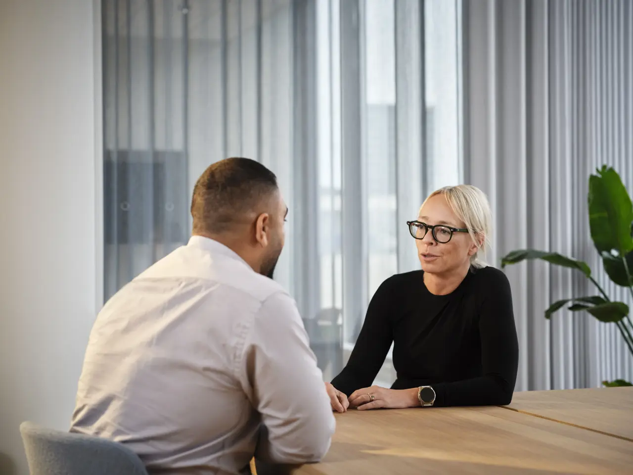 mies ja nainen istuvat pöydän ääressä ja keskustelevat. He ovat neuvotteluhuoneessa. Kasvi taustalla.