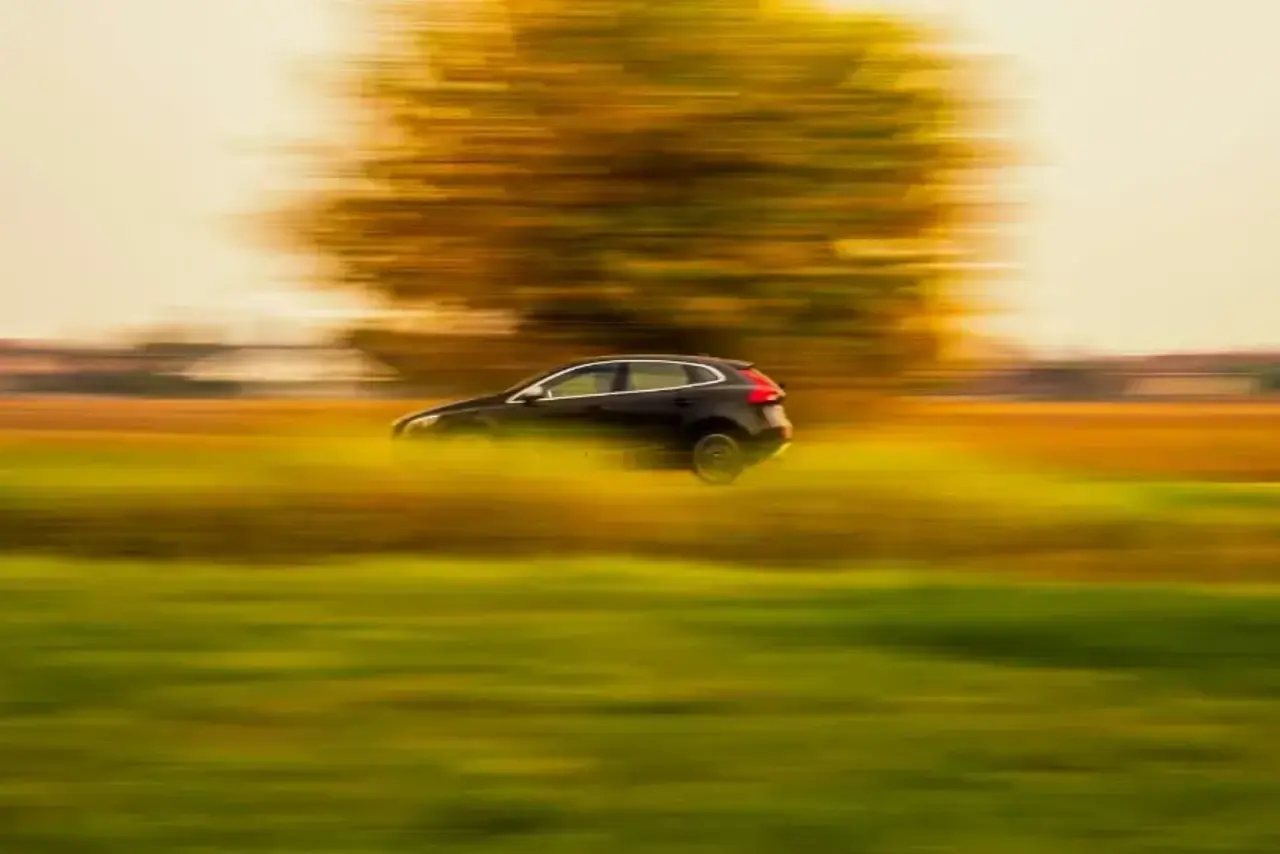 Bild på sidan av en svart bil som kör i hög hastighet. Framför bilen finns en suddig grön platt gräsplätt med lite brunt högre gräs intill bilen. I bakgrunden syns ett syddigt stort träd med bruna och gröna löv. Bakom trädet syns en gräsplätt med endast brunt gräs.