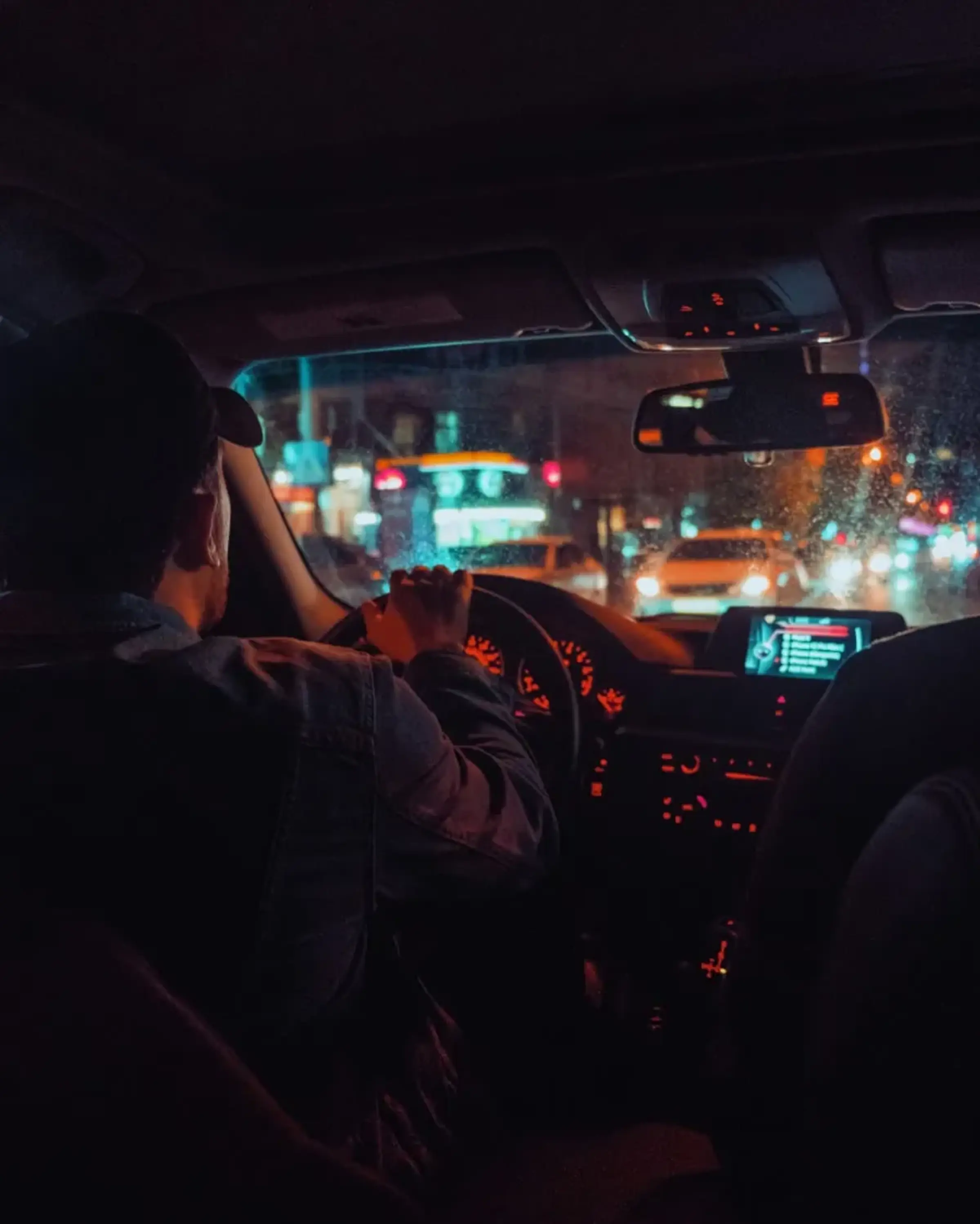 Man i keps kör bil i mörkret. Lamporna inuti bilen lyser rött. Flera bilar kommer mötandes på andra sidan vägen.