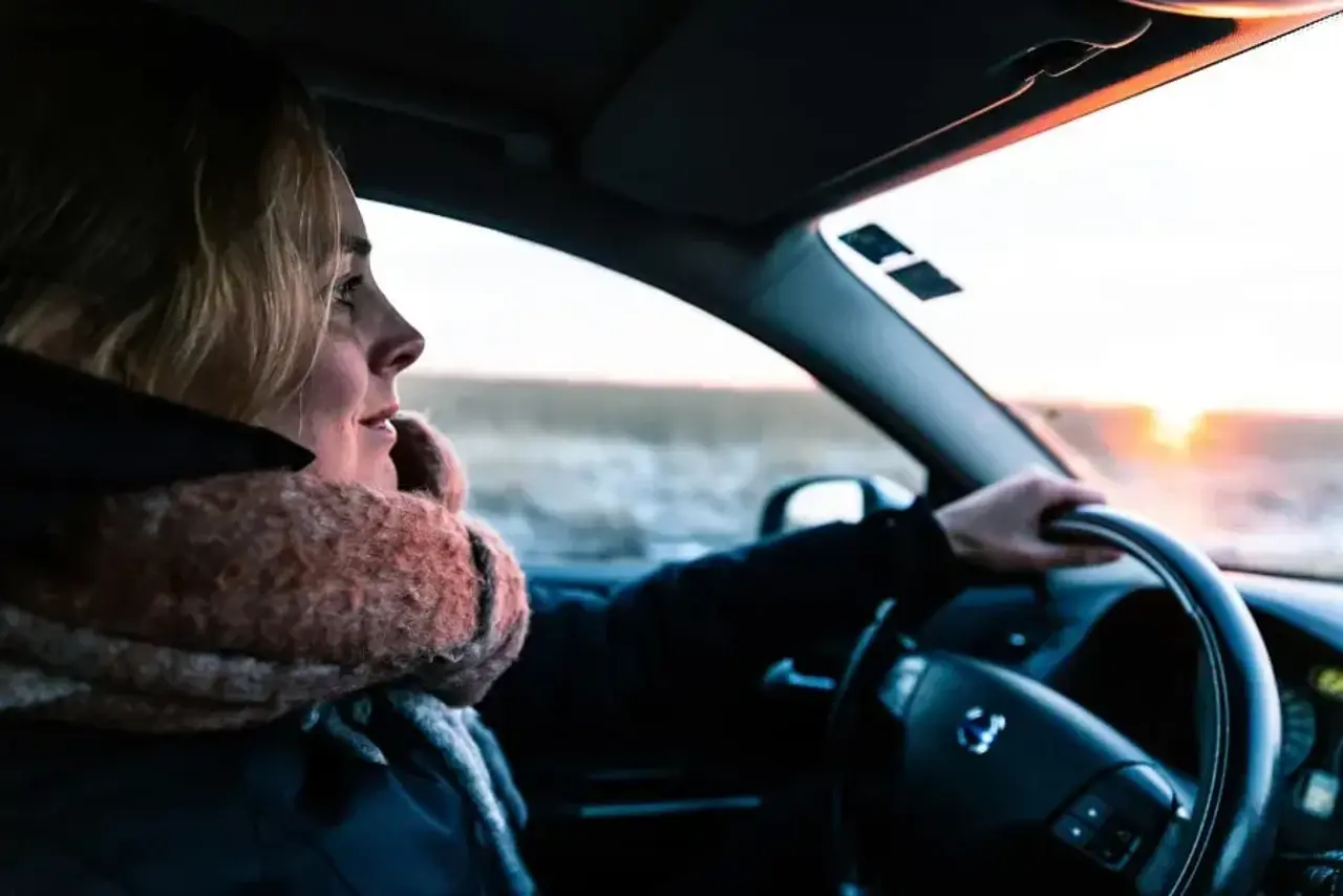 Blond kvinna i vinterjacka och tjock röd halsduk har en hand på ratten och kör bil i soluppgången på vintern. Utanför bilen syns ett fält av högt gräs.
