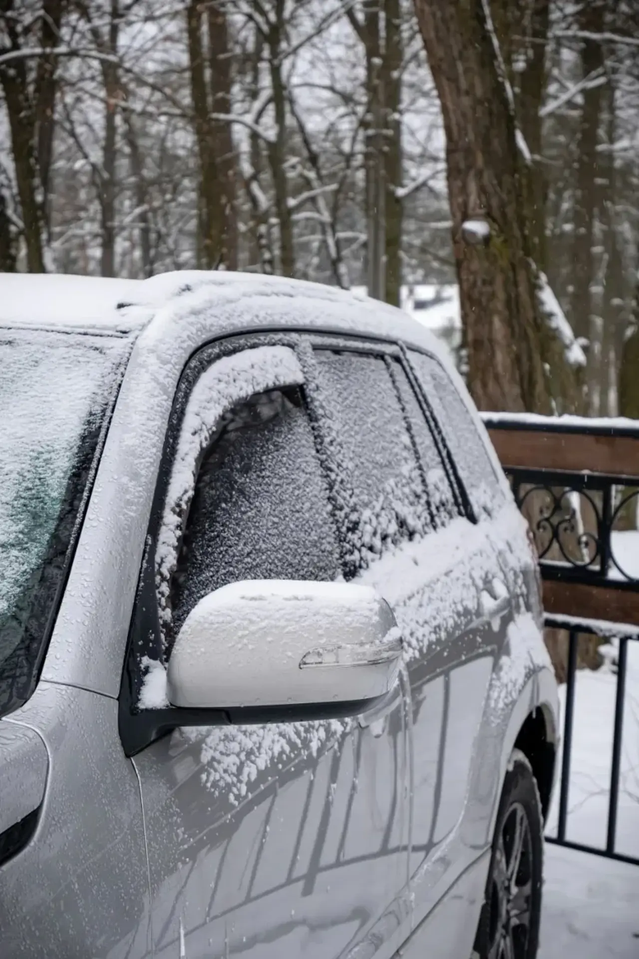 En grå bil står stilla på en snötäckt parkering. Bilen är delvis täckt av snö och frost. I bakgrunden syns flera träd utan löv. Ett staket sträcker sig bakom bilen, delvis täckt av snö. I bakgrunden finns även ett tegelhus.