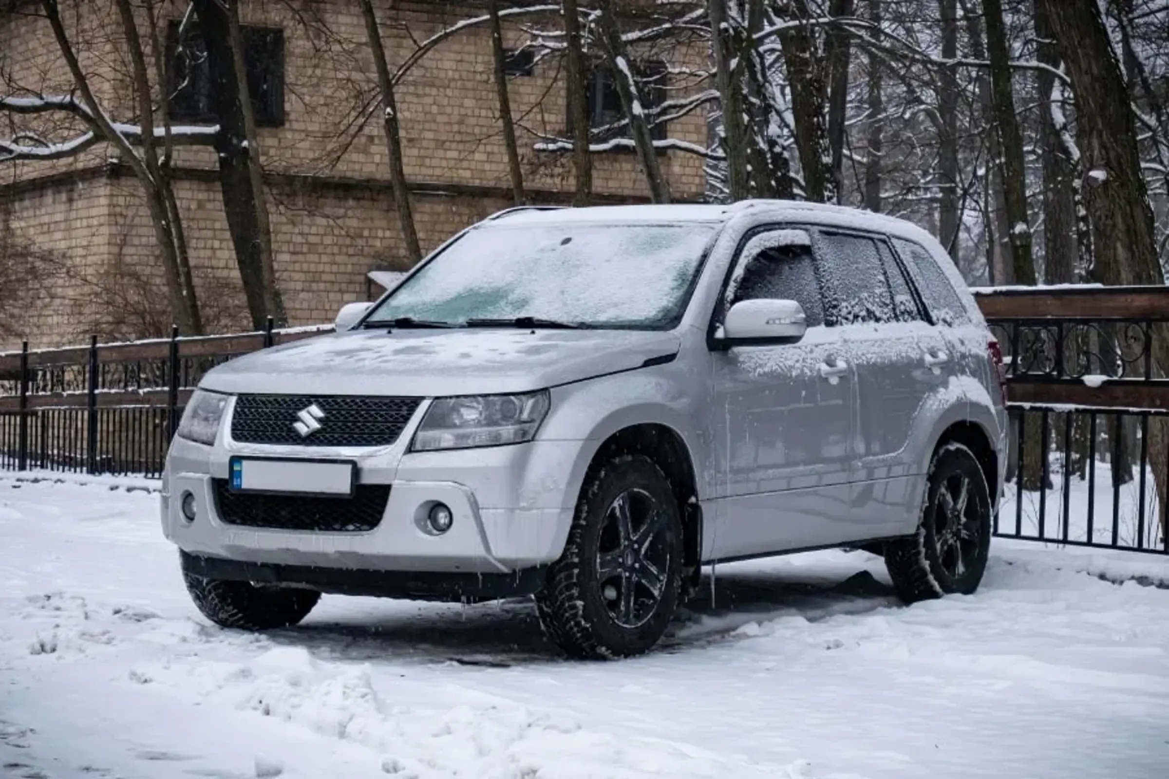 En grå bil står stilla på en snötäckt parkering. Bilen är delvis täckt av snö och frost. I bakgrunden syns flera träd utan löv. Ett staket sträcker sig bakom bilen, delvis täckt av snö. I bakgrunden finns även ett tegelhus.