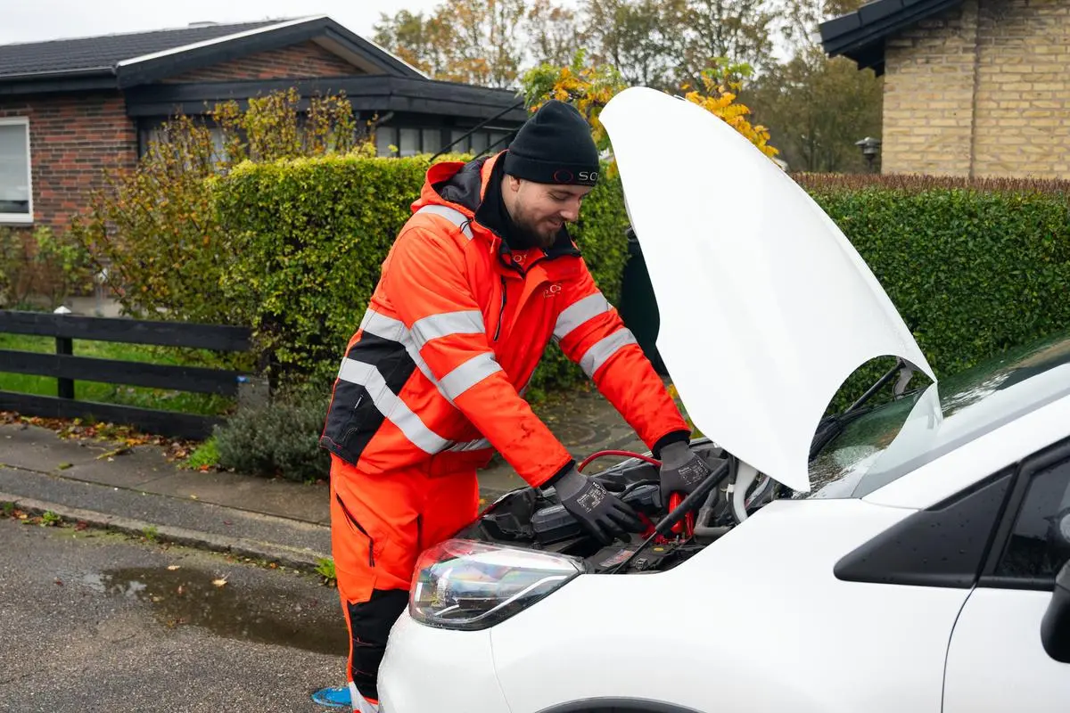 Autoredder ses fra siden stå ved bil. Er i gang med at jumpstarte bilen med booster. 