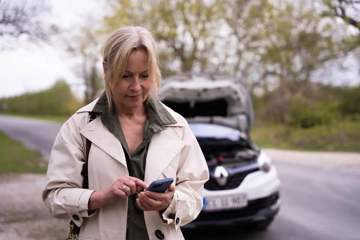 Dame står med telefon i hånden. I baggrunden er bil med motorhjelm oppe.