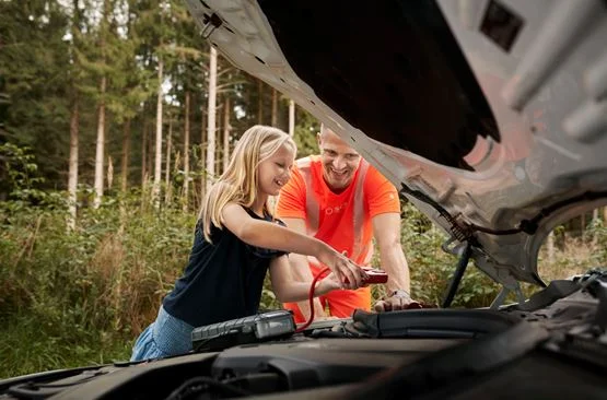 Autoredder viser pige hvordan man sætter en booster på et bilbatteri. Motorhjelm åben. 