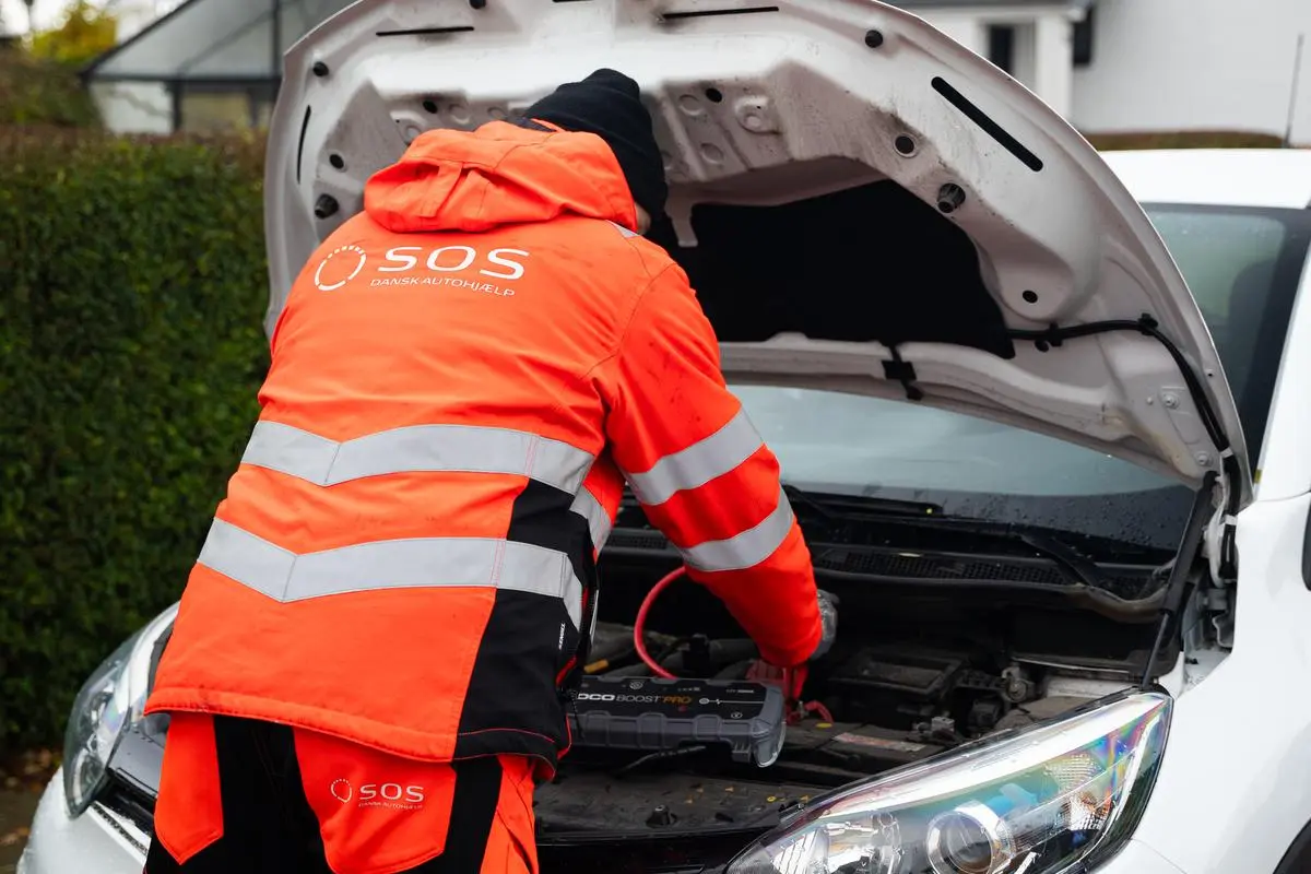 Autoredder står med rykken til og er i gang med at starte bil. Motorhjelm er åben og der er påsat booster til batteriet på bilen. 