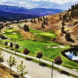 An aerial view of the Black Mountain Golf Club in Kelowna with lush green fairways, sand bunkers, and a winding road in the foreground, set against a backdrop of rolling hills and mountains.