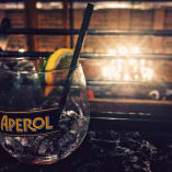 A glass of Aperol with a straw and a lemon slice is placed on a dark surface in BNA Brewing, Kelowna, with blurred lights in the background.