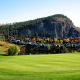 A scenic view of Gallagher's Canyon Pinnacle Course in Kelowna, featuring green fairways, a picturesque pond, homes nestled among vibrant autumn trees, and a rocky hill covered with pine trees in the background.