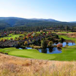 View of Gallagher's Canyon Pinnacle Course in Kelowna featuring green lawns, trees with autumn foliage, houses, ponds, and mountains in the background.