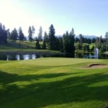 A beautiful golf course with a pond and fountain, surrounded by trees and mountains in the background, at Kelowna Golf & Country Club.