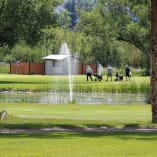 a group of people walking across a lush green field next to a pond with a fountain in the middle of it