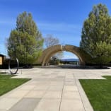 A view of Mission Hill Family Estate Winery in West Kelowna featuring a large archway structure, paved pathway, and manicured lawns with trees under a clear blue sky.
