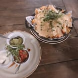 A skillet of cheesy nachos garnished with herbs and a plate of dipping sauces on a wooden table at Rustic Reel Brewing Co. in Kelowna.