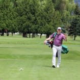 a man walking across a green covered golf course with a bag of golf balls in his hand and a golf ball in his other hand