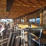 a group of people sitting at tables under a wooden roof overlooking a lake and mountains in the distance