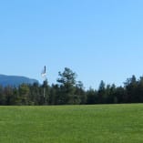 A well-maintained golf course with a visible flagstick set against the backdrop of trees and distant hills under a clear blue sky at Two Eagles Golf Course in West Kelowna.