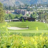 A scenic view of Two Eagles Golf Course in West Kelowna with greenery, a sand bunker, and buildings against a mountainous backdrop.