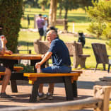 A couple of people sitting at a picnic table in a park with drinks in their hands, one holding a cell phone.