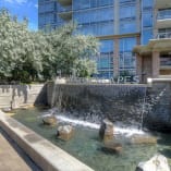 A small waterfall feature at Waterscapes in downtown Kelowna, surrounded by trees and rocks in front of a modern multi-story building.