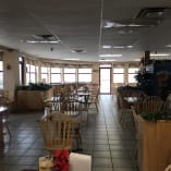 A restaurant with tables, chairs, and potted plants in the center of the room and a bar in the middle of the room.