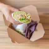 a person holding a burrito in a box on top of a wooden table next to a purple napkin