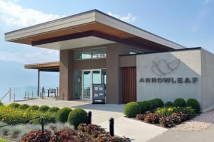 A modern building with Arrowleaf signage, surrounded by greenery and an outdoor seating area overlooking a scenic view at Arrowleaf Cellars in Lake Country.