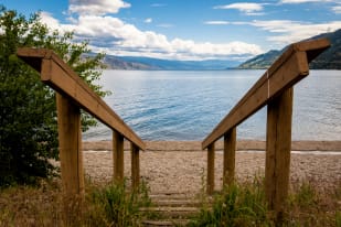 A wooden staircase leads down to the pebbled shore of a serene lake surrounded by mountains under a partly cloudy sky.