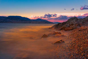 A rocky shoreline with water reflecting the vivid colors of a dramatic sunset over the mountains in Kelowna.