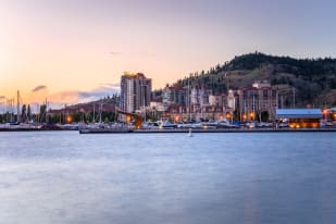 A waterfront view in Kelowna at sunset with boats docked, buildings, and a hillside in the background.