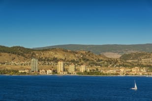 A view of Kelowna with buildings and hills in the background, and a sailboat on the water in the foreground.