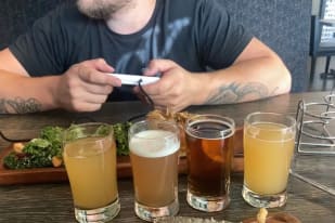 A man sitting at a table with four glasses of beer, a cell phone, and a plate of food in front of him.
