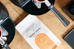 A coffee maker on a wooden table next to a bag labeled Bright Jenny Coffee Roasters and a cup filled with coffee grounds.