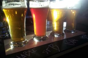 A flight of four craft beers in different colors served in glasses on a wooden paddle at The Curious Artistry & Alchemy Cafe in Kelowna.