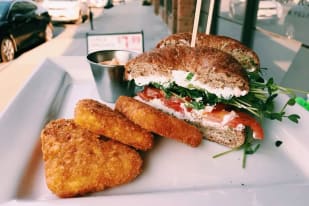 A close-up of a sandwich with fresh vegetables on a bagel, accompanied by crispy fried potato patties, served at an outdoor cafe.