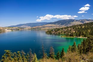 Scenic view of a tranquil lake surrounded by lush greenery and mountains in Vernon, under a clear blue sky with scattered clouds.