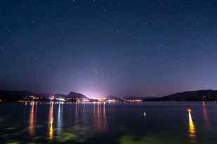 The image features a serene nighttime scene with a large body of water, a star-filled sky, and a cityscape in the background in Okanagan.