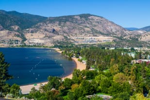 A scenic view of Penticton with a sandy beach along a blue lake, surrounded by greenery and mountains in the background.