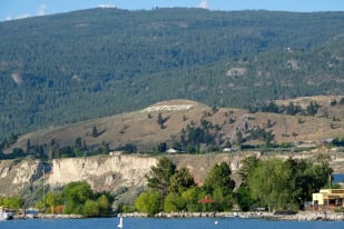 Hillside with PENTICTON written, seen across a body of water with trees and buildings in the foreground.