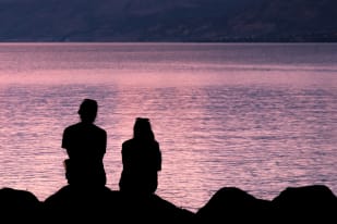 Two silhouetted people sit on rocks, gazing at a serene lake during sunset in Penticton.