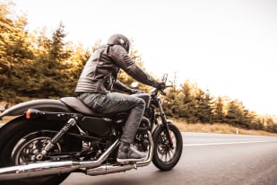 A person in a black leather jacket riding a motorcycle on a road with trees in the background.