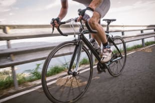 A cyclist riding a road bike along a scenic route with a metal guardrail and water view in the background.