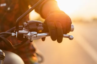 A gloved hand gripping the handlebar and brake lever of a motorcycle at sunset.