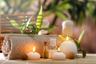 A serene spa setup with lit candles, essential oil bottles, and a rolled towel on a bamboo mat surrounded by green plants.