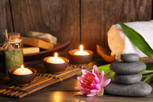 Spa scene with lit candles, stacked stones, a pink flower, soap bars, and a rolled white towel on a wooden surface.
