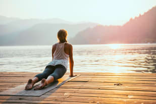 A woman is practicing yoga in an upward-facing dog pose on a wooden dock, facing the serene water during sunset.