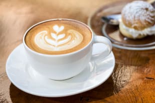 A cup of cappuccino with latte art on a white saucer beside a cream puff pastry on a glass dish.
