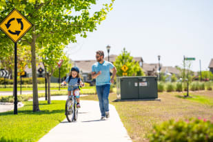 A father jogs alongside his young daughter as she rides her bike on a sunny neighborhood path with trees and a roundabout sign nearby.